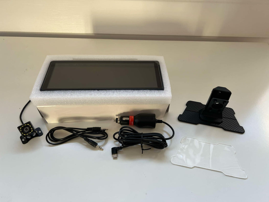 CarPlay-skjerm med ryggekamera, strømadapter, kabler og dashbordfeste lagt frem på et bord.